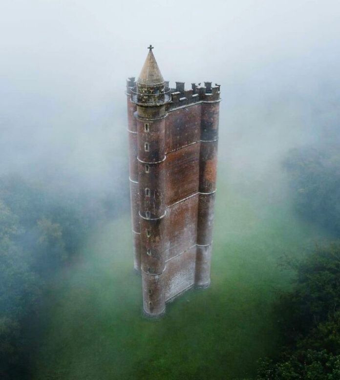 Fog-shrouded red-brick tower with a pointed roof and battlements, rising vertically from green grass into misty air, creating an isolated, fantasy-like atmosphere.