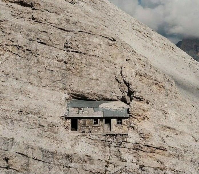 Camouflaged wooden cabin embedded into a rocky mountain cliff in the Italian Alps, blending almost invisibly with the stone surroundings.