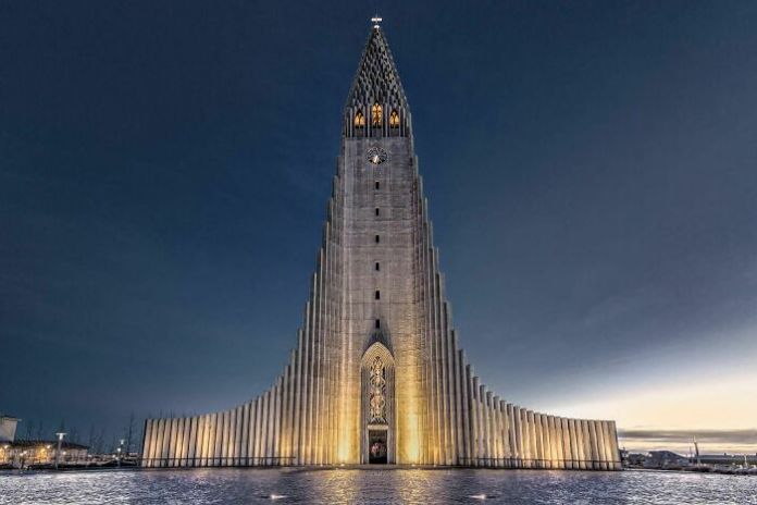 Symmetrical concrete church in Iceland with a soaring tower and stepped wings, glowing with warm lights against a deep blue evening sky.