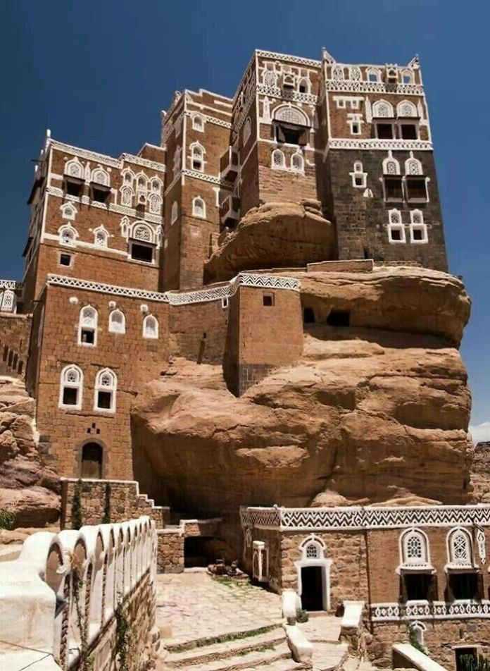 Multi-story brown stone buildings in Sanaa, Yemen, with white geometric window detailing, built atop and integrated with natural rock formations.