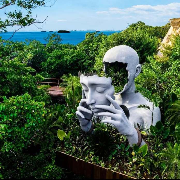 Large white sculpture in a tropical garden by Daniel Popper, depicting a human figure holding its own facial mask in both hands, with the top of the head open and filled with lush green plants, set against a backdrop of forest and ocean in Cancun, Mexico.