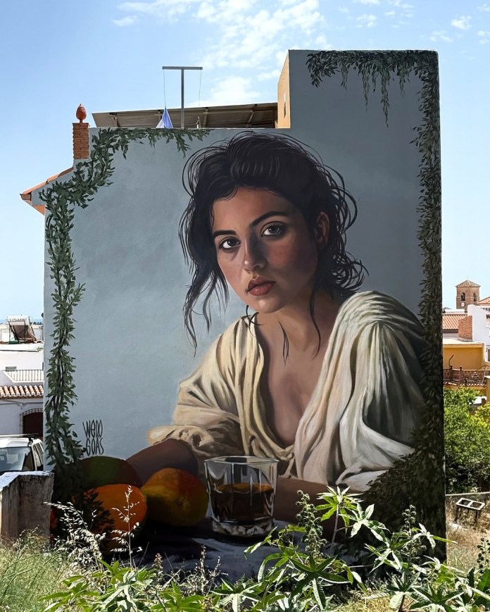 Photorealistic mural on a building of a dark-haired woman in a cream shirt, leaning on her arm, with mangoes and a glass of drink on the table in front of her, framed by painted green vines.