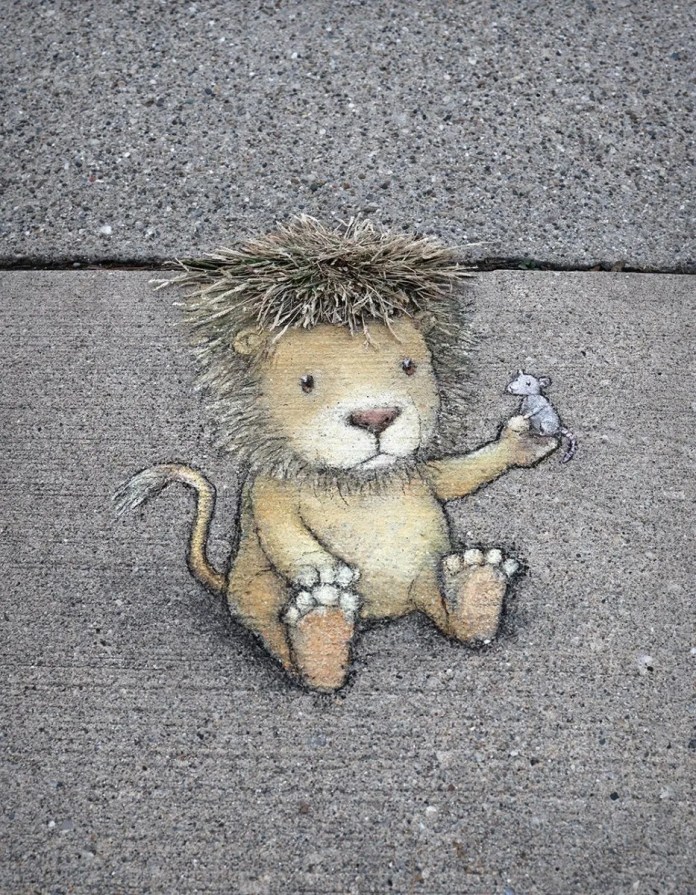 A whimsical chalk drawing by street artist David Zinn on a concrete sidewalk, featuring a small lion with soft golden fur, a playful expression, and a mane creatively integrated with real grass. The lion is sitting down, holding a tiny gray mouse in one paw, evoking the classic fable of "The Lion and the Mouse." The artwork seamlessly blends with the urban environment, using natural elements to enhance its charm and humor.
