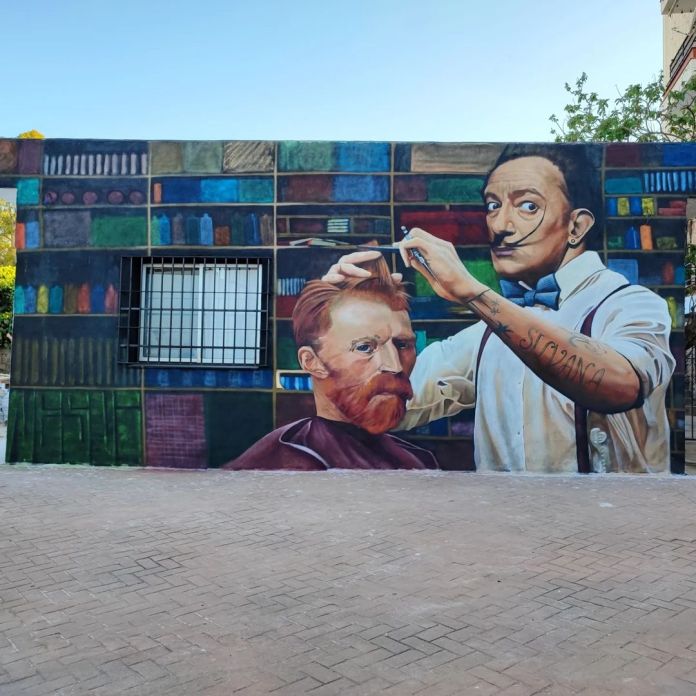 A vibrant street art mural in Malaga, Spain, depicting Salvador Dalí styling Vincent Van Gogh's hair in a surreal barber scene. Painted by artist Nesui, the artwork shows Dalí holding scissors with a playful expression, while Van Gogh sits in a barber's chair with a calm demeanor. The backdrop features shelves filled with colorful books and objects, blending playful elements with homage to these iconic artists.