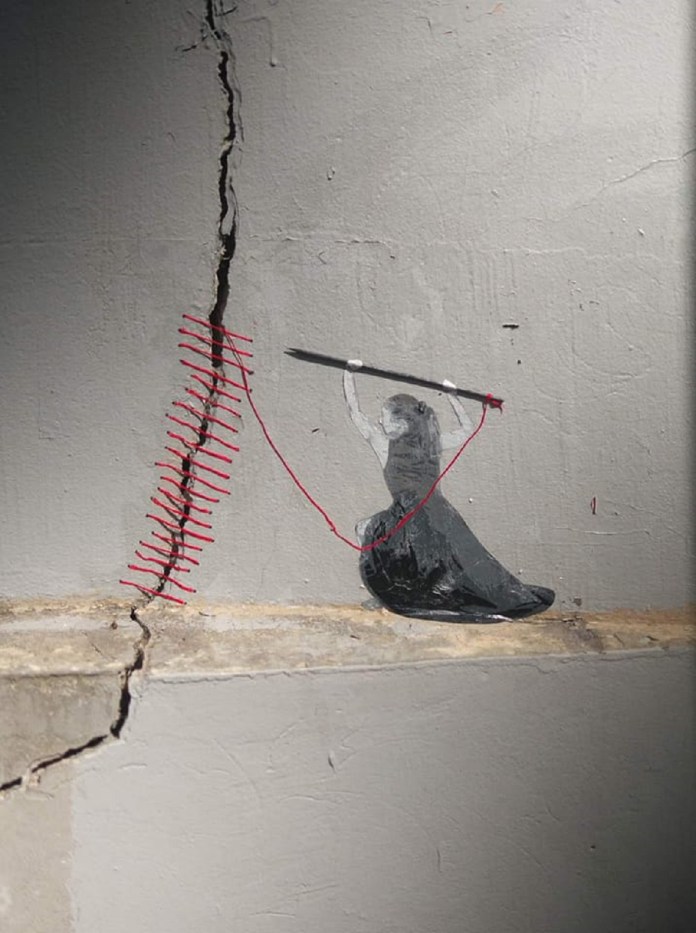 Mural of a female figure at the base of a cracked wall, pulling red thread through both sides to stitch the damage closed, blending 2D paint with real red thread.