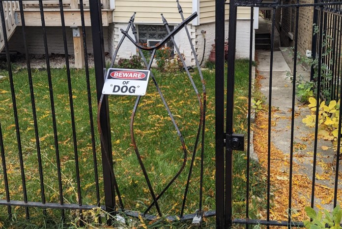 Metal gate with warped bars forming the shape of a dog mid-leap through an opening, accompanied by a “Beware of Dog” sign hanging askew.