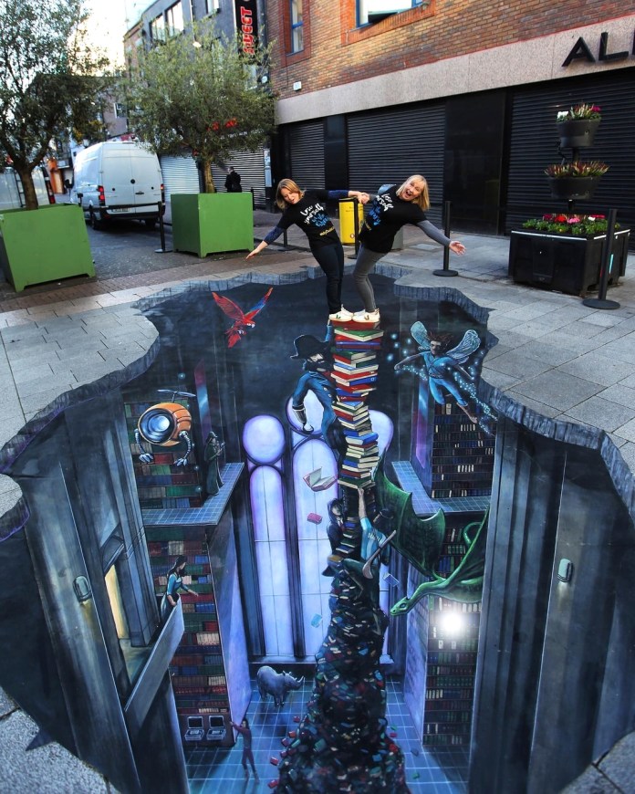 3D street art titled “Tower of Stories” in Dublin, Ireland, showing an imagined underground library with a spiraling tower of books surrounded by fantasy figures like dragons, astronauts, and flying readers.