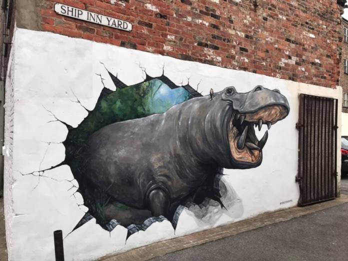 Mural titled “Hippo Breakout” showing a roaring hippopotamus crashing through a painted wall with broken edges and exposed brick surrounding the scene.