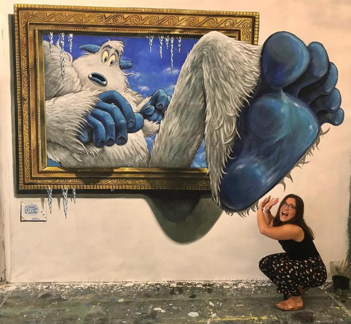 3D mural by Joe and Max, showing a white-furred yeti with blue hands and feet stepping out of a golden picture frame with icicles, as a woman crouches below playfully acting surprised.