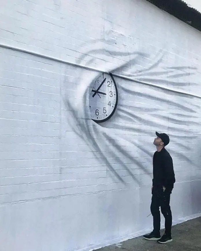 A black-and-white mural featuring a hand stretching what looks like fabric around a real clock, giving the illusion that time is being physically pulled. A man in black stands below, looking up.