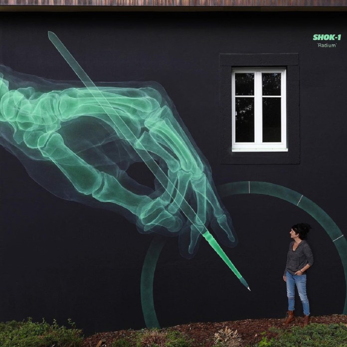 A large green X-ray-style mural of a skeletal hand holding a pencil, which is drawing a glowing arc resembling a clock face. Painted on a black wall with a real window in the scene.