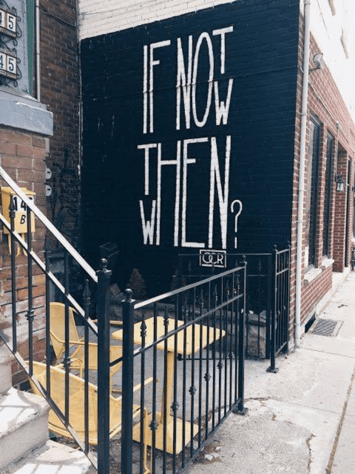 A mural painted in tall white letters on a black brick wall reads “IF NOT NOW THEN WHEN?” with the word “WHEN” vertically stylized to fit narrow space.