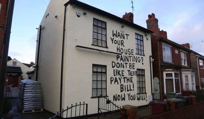 Black text painted in capital letters on the side of a white house says, “WANT YOUR HOUSE PAINTING? DON’T BE LIKE TERRY. PAY THE BILL! NOW YOU WILL!”