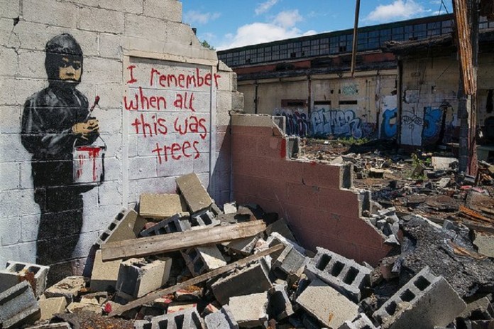 A poignant piece by Banksy located in Detroit, USA. This artwork features a young boy holding a paintbrush and a dripping red paint bucket, standing next to the graffiti text: "I remember when all this was trees." The mural is painted on a crumbling wall amidst rubble and urban decay, evoking themes of deforestation, industrialization, and environmental loss. The background shows an abandoned structure adorned with graffiti, amplifying the message of neglect and ecological destruction.