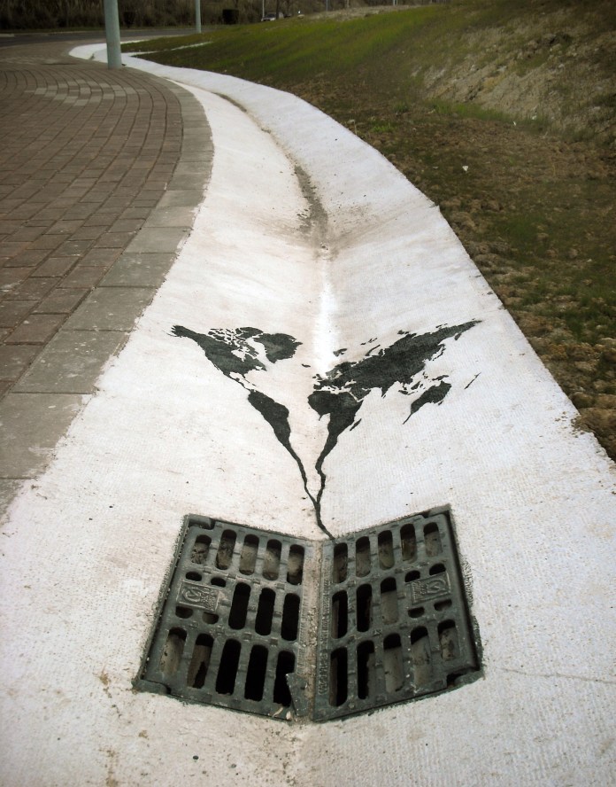 A striking street art piece by Pejac located in Santander, Spain. This artwork features a painted map of the world seemingly flowing into a storm drain, symbolizing environmental degradation and the loss of natural resources. The minimalist design, placed on a white concrete surface along a roadside, uses the shape of the drain to emphasize the visual metaphor of Earth's decline. The piece invites reflection on sustainability and our impact on the planet.