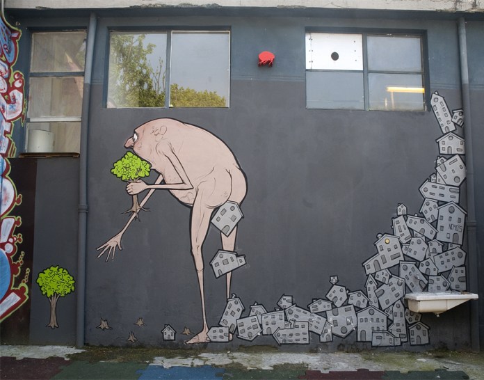 A thought-provoking mural titled "Cagacemento" by NemO’s, located in Milan, Italy. The artwork features a naked, hunched humanoid figure with elongated limbs, clutching a small green tree. Behind the figure, a trail of gray, block-like houses spills out from its body, symbolizing urban sprawl and environmental destruction. In contrast, tiny vibrant trees grow at the edges of the mural, emphasizing the conflict between nature and relentless human expansion. The stark, minimalist design on a gray wall underscores the ecological message of preserving greenery amidst urbanization.