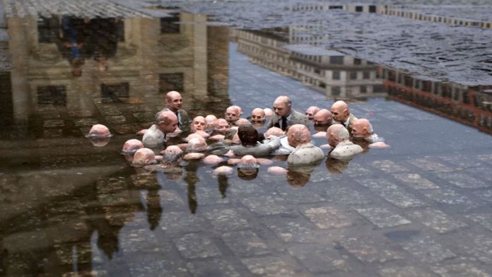 A powerful installation by Isaac Cordal titled "Follow the Leaders," located in Berlin, Germany. This piece features small, detailed sculptures of suited businessmen standing in a flooded urban setting, with only their heads and shoulders visible above water. The figures appear engaged in a discussion, oblivious to their dire surroundings, symbolizing political inaction and corporate greed amidst the climate crisis. The reflective water amplifies the somber tone, while the urban backdrop emphasizes the disconnect between decision-makers and environmental realities.