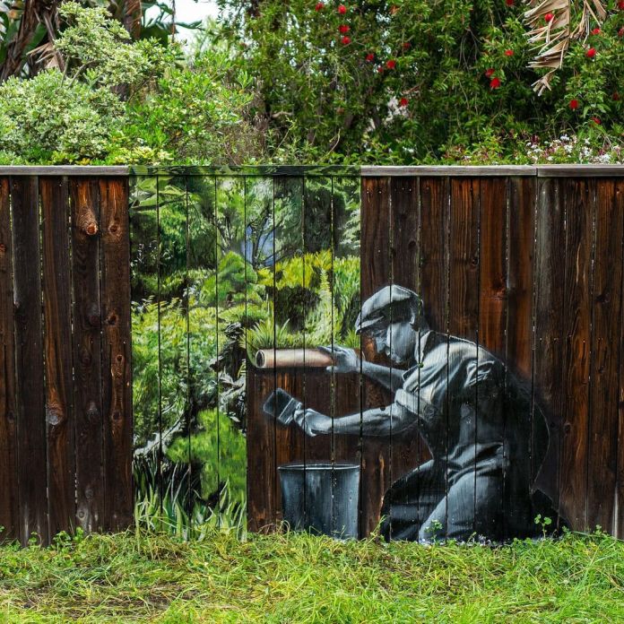 A mural by street artist HIJACK titled "Make Earth Green Again," painted on a wooden fence. The artwork depicts a man wearing a hat and kneeling as he carefully applies green paint to the natural surroundings. The fence itself is seamlessly integrated into the piece, with the wooden slats blending into the painted greenery beyond, creating the illusion of a continuous forest. The message underscores the importance of restoring and protecting nature amidst urban environments. The vibrant greenery contrasts beautifully with the dark wood, drawing attention to the theme of ecological renewal.