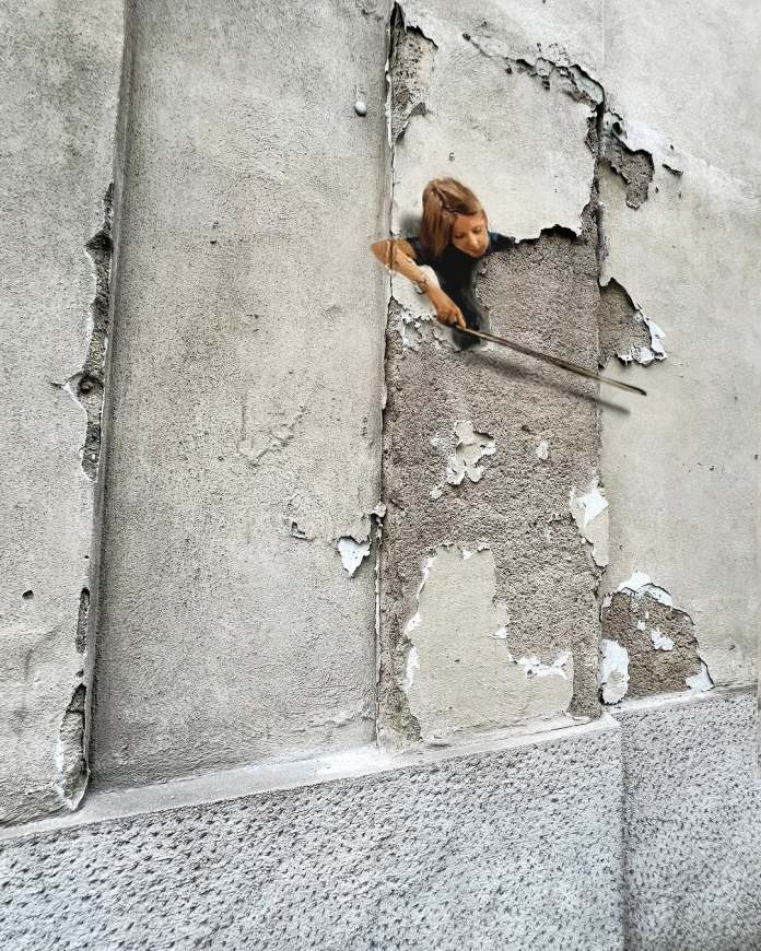 Street art in Milan by Golsa Golchini depicting a girl holding a bow, appearing to play a Double Bass made from the peeling plaster and cracks on the wall. The illusion transforms the broken surface into a poetic urban instrument.