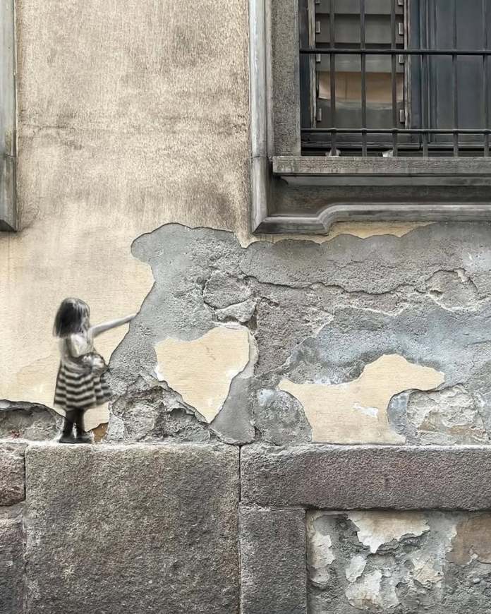 Street art in Milan by Golsa Golchini showing a young girl in a striped dress reaching out to pet an elephant, with the animal’s shape created from peeling paint and rough wall textures. The scene creates a tender interaction using only minimal painted elements and surface damage.
