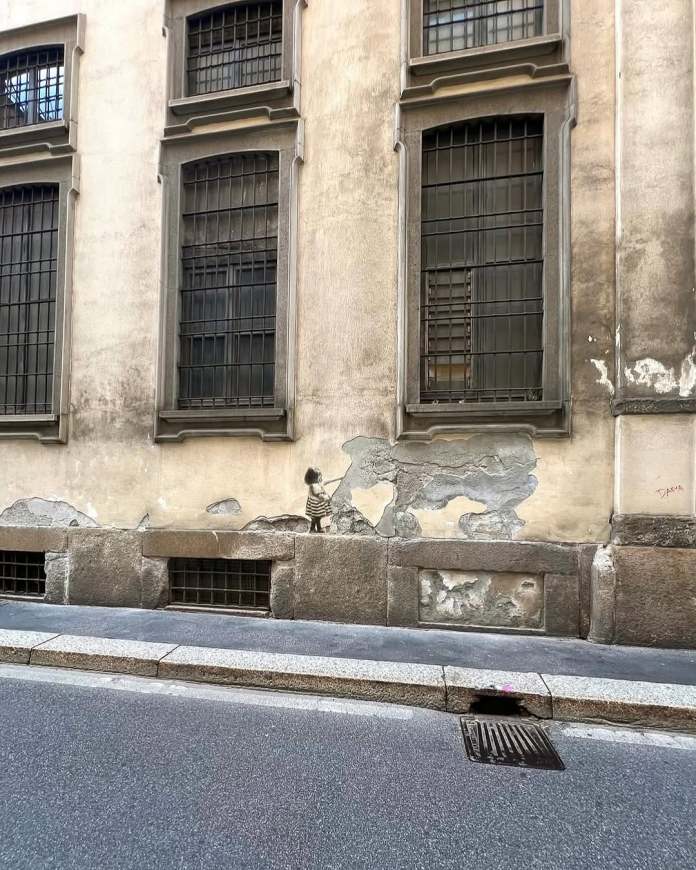 Street art in Milan by Golsa Golchini showing a young girl in a striped dress reaching out to pet an elephant, with the animal’s shape created from peeling paint and rough wall textures. The scene creates a tender interaction using only minimal painted elements and surface damage.