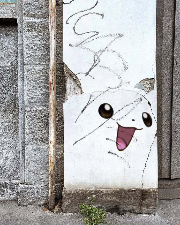 Street art in Milan by Golsa Golchini featuring a Pikachu-inspired face painted onto a cracked wall. The design uses large anime-style eyes and an open pink mouth, with peeling plaster integrated into the expression.