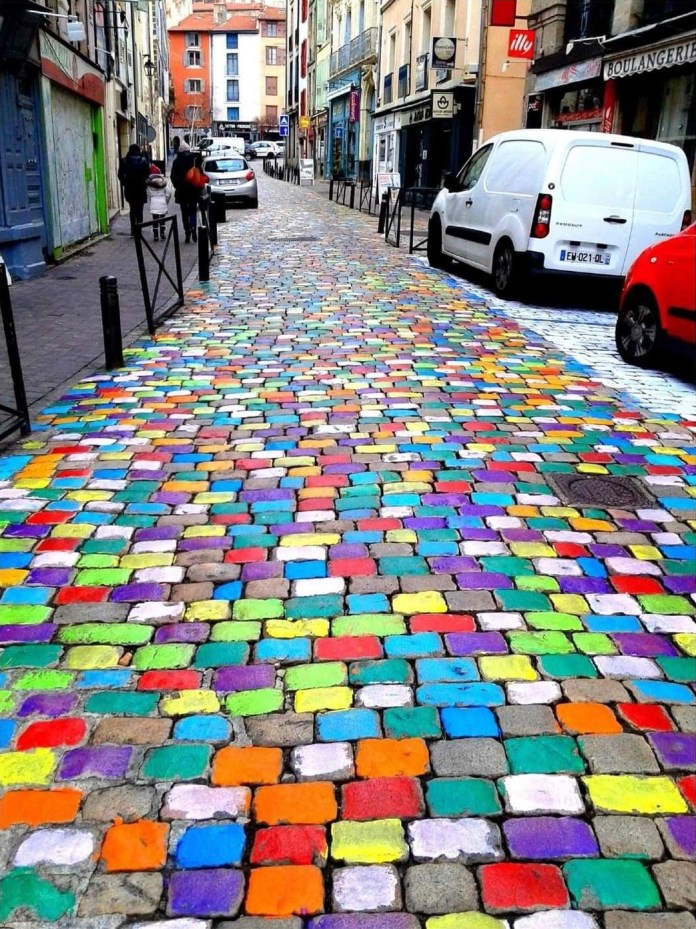 Painted cobblestone street in Le Puy-en-Velay, France, with each stone coated in bold colors such as red, green, yellow, blue, and purple, creating a rainbow effect down the road.