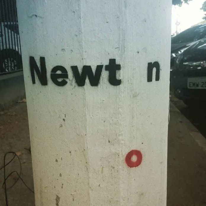 Street art on a white pole spelling “Newt n” with a red painted apple on the ground, visually completing the word “Newton.”
