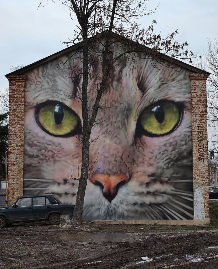 Large mural by LexusOne in Nizhny Novgorod, Russia, depicting a close-up of a cat’s face with bright green eyes painted across the entire wall of a brick building.