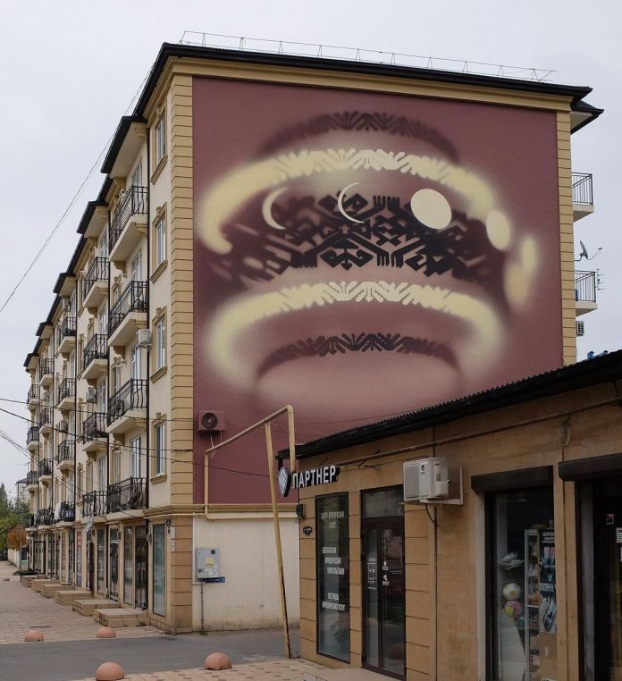 Street art mural by Shozy in Derbent, Russia, featuring an abstract, symmetrical design resembling traditional mountain patterns, glowing with circular and crescent shapes on a soft maroon wall.