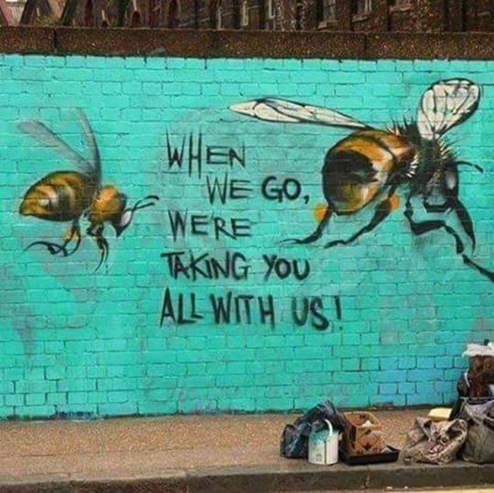 Mural by Louis Masai featuring two large bees painted on a turquoise brick wall with the handwritten message “When we go, we’re taking you all with us,” located at Shoreditch Station in London, UK.