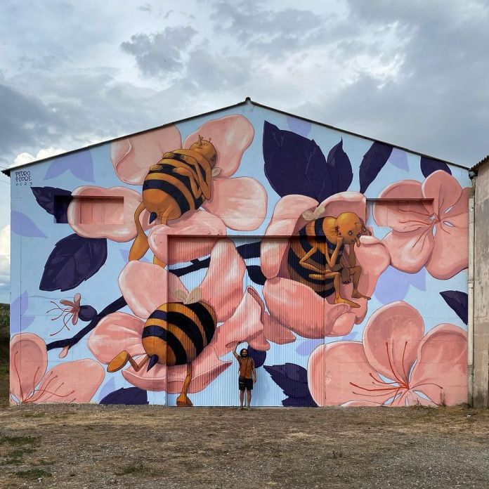 Mural covering a full barn-like building with large pink flowers and three bees featuring childlike human heads, surrounded by purple leaves on a cloudy day.