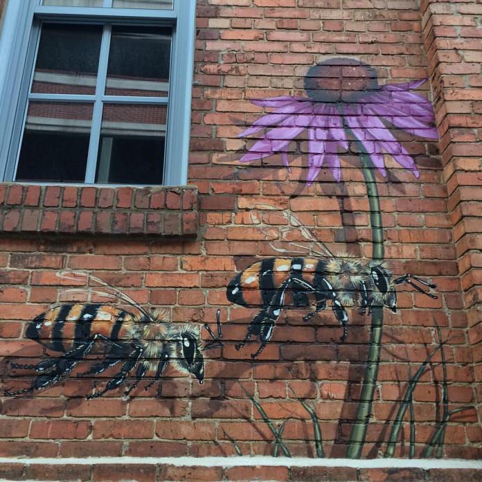Detailed mural of two realistic bees flying near a purple coneflower on a red brick wall beneath a window, part of The Good of the Hive project.