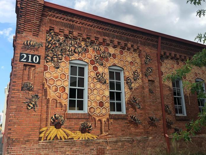 Mural on a red brick building in Durham, North Carolina, featuring dozens of realistic bees crawling across a detailed honeycomb pattern with golden honey dripping down. Yellow coneflowers appear at the bottom of the wall, and the windows are seamlessly integrated into the artwork. Created by Matt Willey as part of The Good of the Hive project.