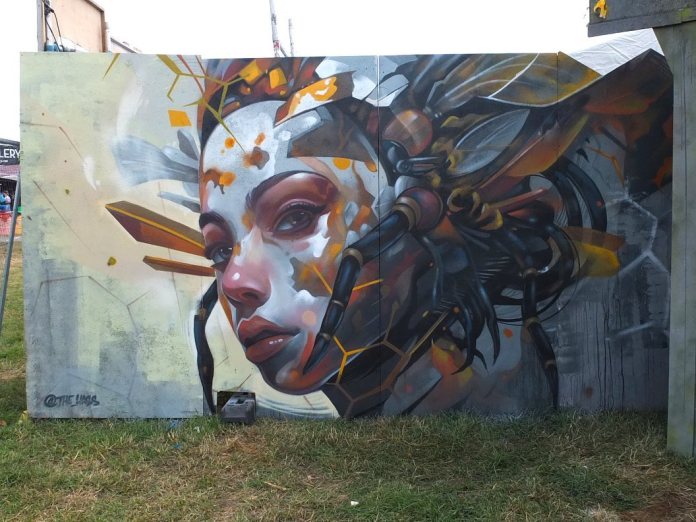Large portrait mural of a woman’s face fused with robotic bee parts, painted in cool tones with orange hexagonal accents and mechanical limbs.