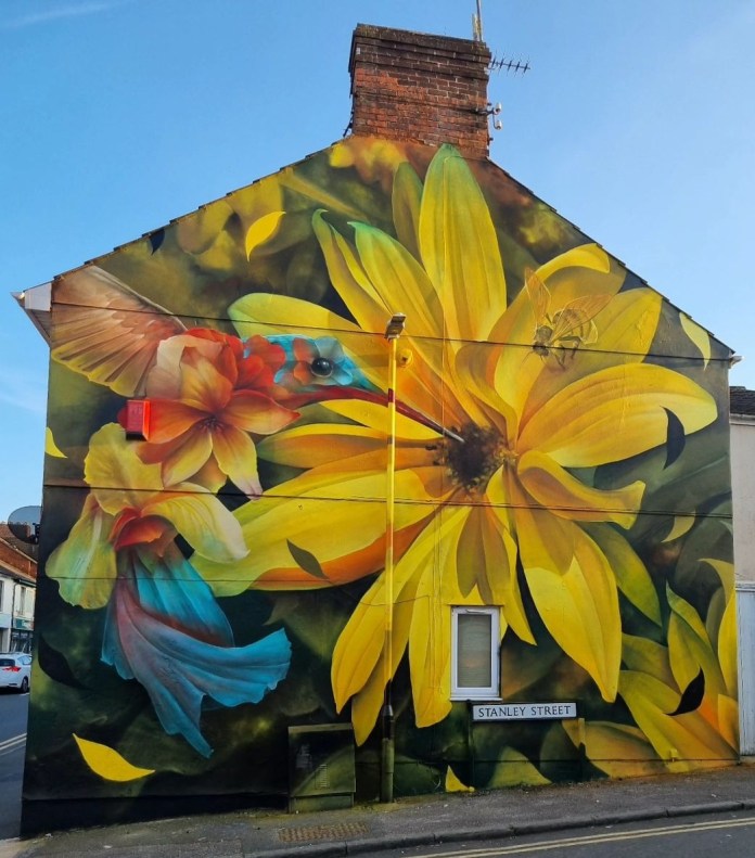 Bright mural on the side of a house showing a hummingbird and a bee approaching a giant yellow flower, painted in photorealistic style.