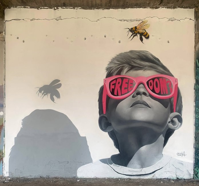 Black-and-white mural of a boy wearing red sunglasses with the word “FREEDOM?” painted across the lenses, looking up at a flying bee on a white wall.