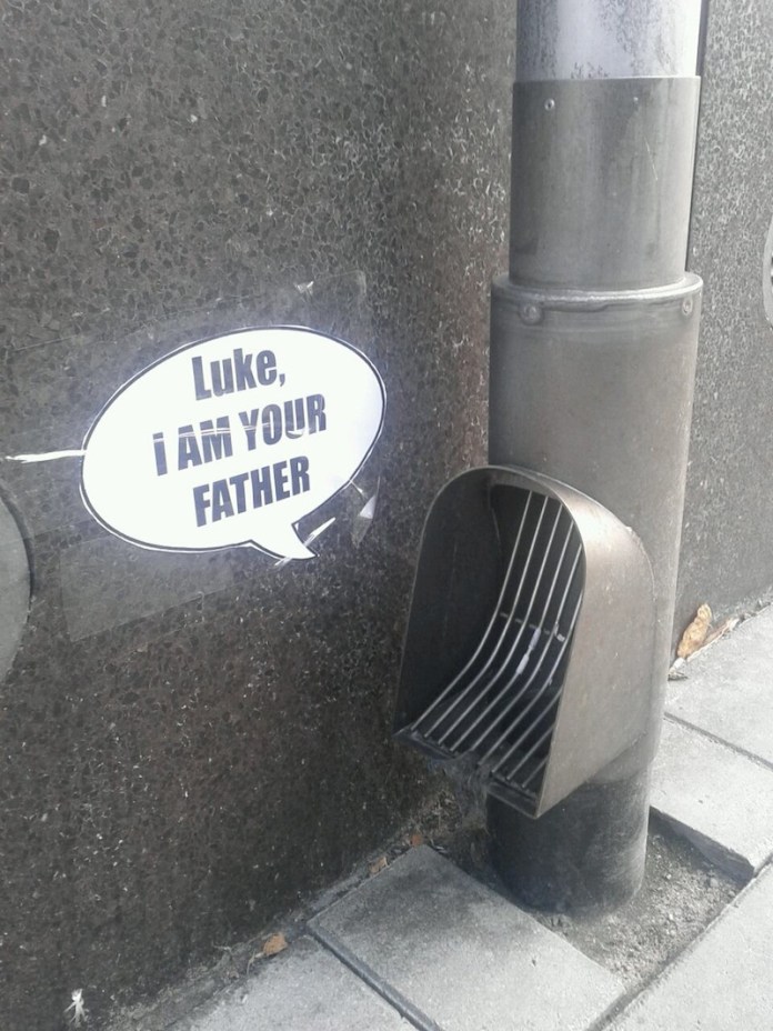 A ventilation pipe on a street wall next to a speech bubble sticker that says “Luke, I am your father,” making the pipe resemble Darth Vader.