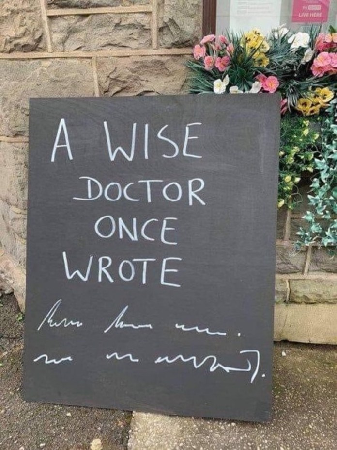 A black sandwich board with the handwritten message "A WISE DOCTOR ONCE WROTE" followed by two lines of intentionally messy scribbles meant to mimic illegible doctor handwriting, placed beside a stone building and flower box.