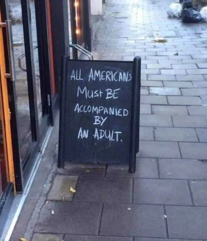 A chalkboard standing on a city sidewalk outside a shop entrance, reading "All Americans must be accompanied by an adult" in uneven handwritten lettering.