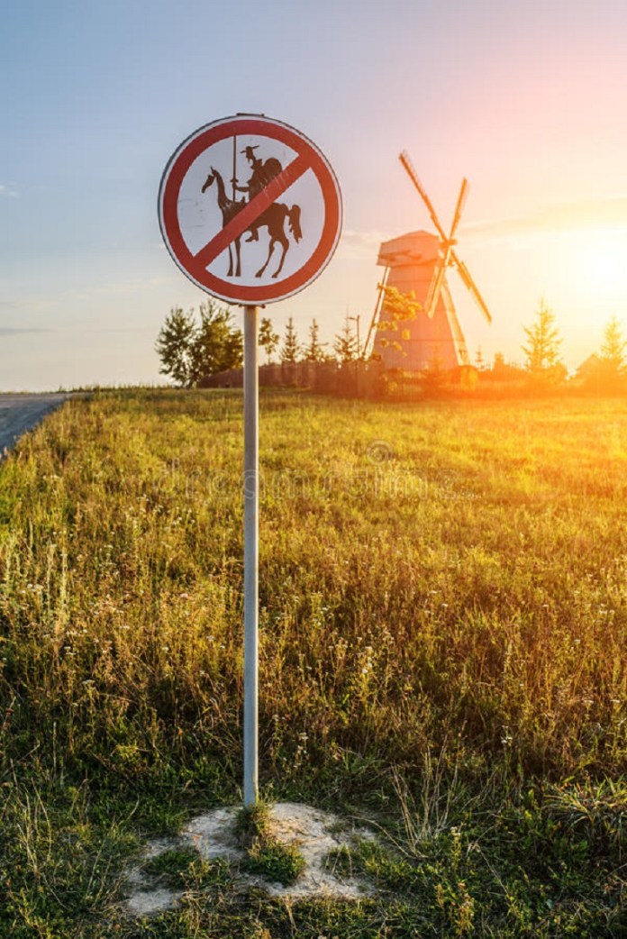 A round road sign with a red diagonal strike banning the silhouettes of Don Quixote and Sancho Panza on horseback, placed in front of a windmill in a grassy field at sunset.