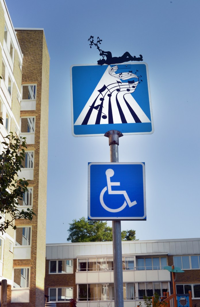 A blue pedestrian crossing sign modified so the crosswalk stripes resemble piano keys turning into music notes, with a silhouette of a dancer added to the top of the sign.
