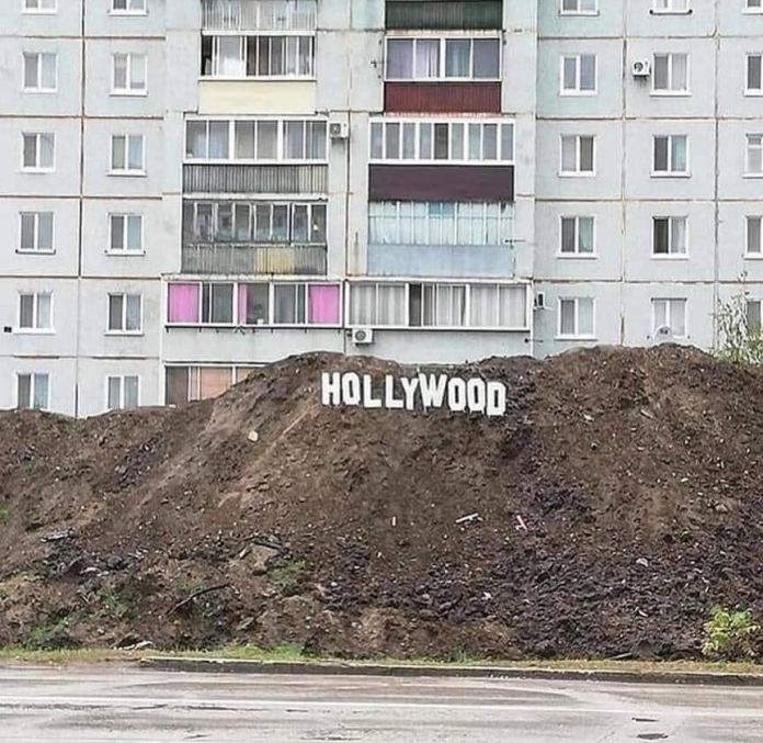 A dirt mound in front of a grey apartment building with white block letters spelling “HOLLYWOOD” attached, mimicking the famous Hollywood sign in a rundown setting.
