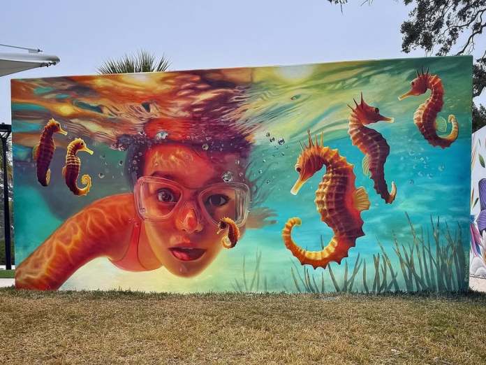 A stunning 3D mural titled "Clear Water Wonders" by street artist Naomi Haverland in Clearwater, Florida, created for the City of Clearwater. The artwork features a young girl with vivid red goggles, partially submerged underwater, gazing directly at the viewer. Surrounding her is a vibrant scene of orange and yellow seahorses floating in clear turquoise water, accentuated by shimmering light effects and aquatic details like bubbles and underwater vegetation. The piece captures a sense of curiosity and connection to marine life.