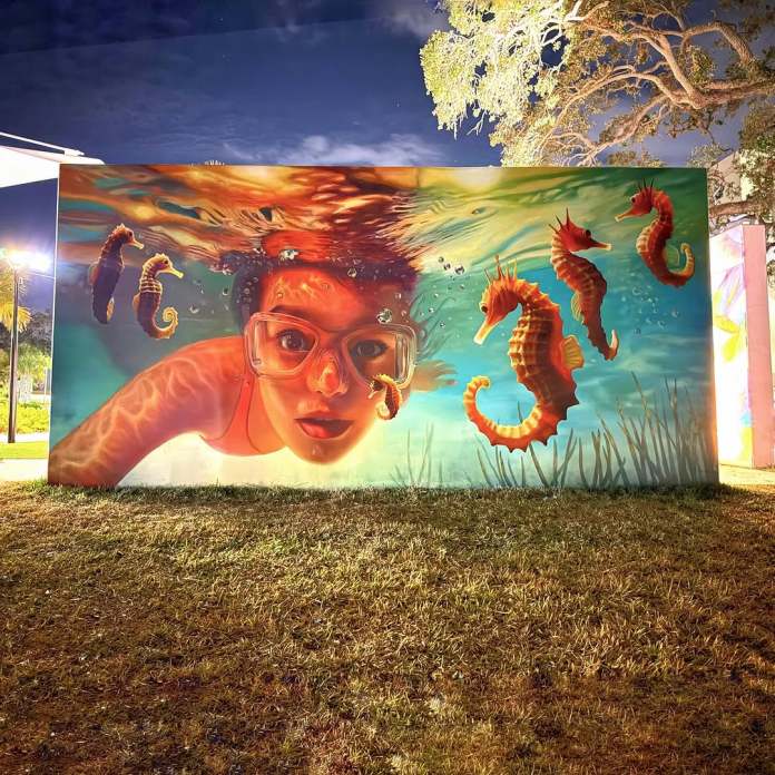 A nighttime view of the 3D mural "Clear Water Wonders" by Naomi Haverland in Clearwater, Florida, created for the City of Clearwater. The artwork depicts a young girl wearing red goggles, partially submerged underwater, with her face illuminated by soft artificial light. Vibrant orange and yellow seahorses appear to float around her in the clear turquoise water, enhanced by detailed light reflections and underwater elements like bubbles and plants. The mural's vivid colors and dynamic composition are beautifully highlighted under the night sky, framed by a large, illuminated tree in the background.