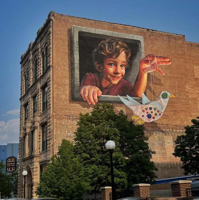 A whimsical 3D mural titled "Hope is a Thing with Sequins" by Naomi Haverland in Sioux City, Iowa. The artwork features a young boy with curly hair, leaning out of a painted window frame on the side of a brick building, holding a string attached to a colorful, handcrafted paper bird. The bird is adorned with vibrant sequins, buttons, and patchwork patterns, symbolizing creativity and hope. The mural blends realism and fantasy, creating a charming and interactive scene against the backdrop of a historic building surrounded by trees and streetlights.