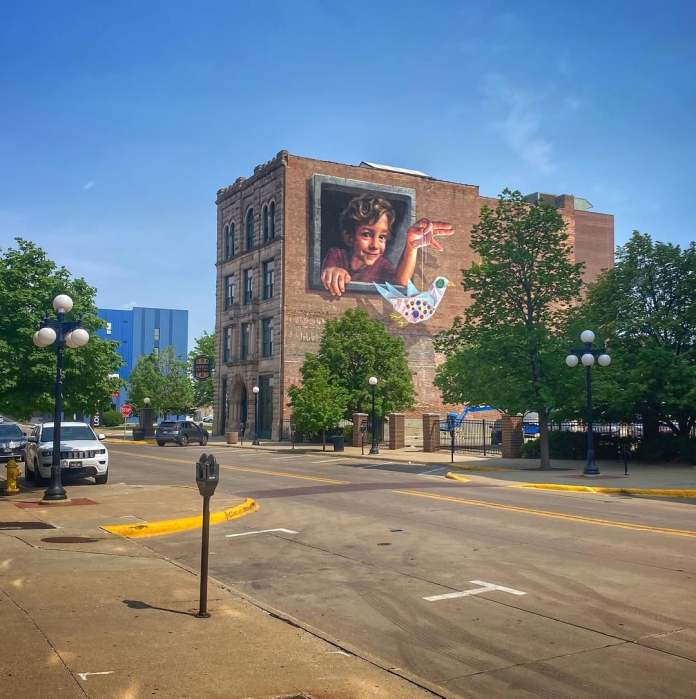 A wide-angle view of the mural "Hope is a Thing with Sequins" by Naomi Haverland in Sioux City, Iowa, located on the side of a historic brick building. The mural depicts a boy leaning out of a painted window frame, holding a string attached to a colorful paper bird decorated with sequins and buttons. The scene blends seamlessly with the surroundings, showcasing the artwork's scale against the urban backdrop, with parked cars, trees, and classic streetlights lining the quiet street under a clear blue sky.