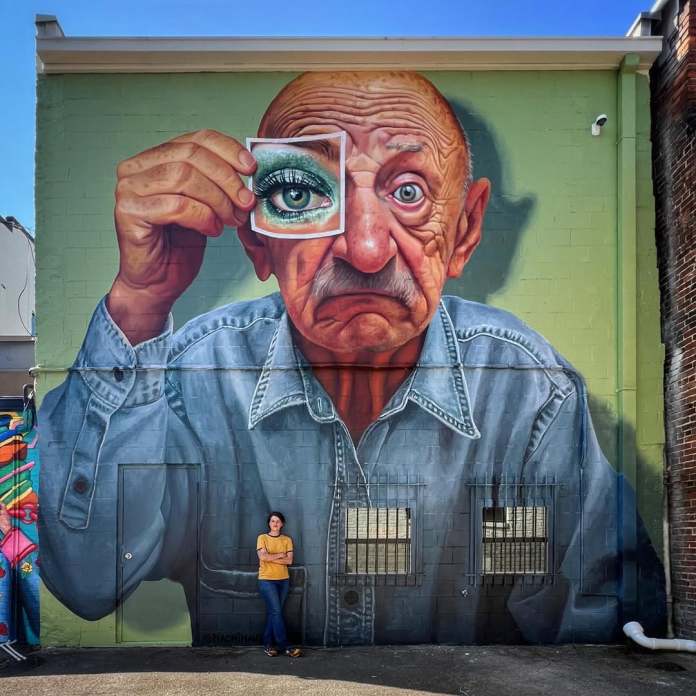 A wide-angle view of the mural "Hope is a Thing with Sequins" by Naomi Haverland in Sioux City, Iowa, located on the side of a historic brick building. The mural depicts a boy leaning out of a painted window frame, holding a string attached to a colorful paper bird decorated with sequins and buttons. The scene blends seamlessly with the surroundings, showcasing the artwork's scale against the urban backdrop, with parked cars, trees, and classic streetlights lining the quiet street under a clear blue sky.