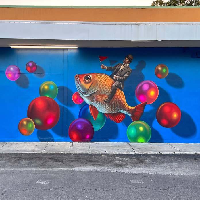 A vibrant mural by Naomi Haverland created for the Art Oasis Mural Fest in Clearwater, Florida. The artwork features a whimsical scene of a man dressed in a suit and top hat, holding a small red flag as he rides a large, colorful fish. The fish is surrounded by glowing, multicolored bubbles that seem to float against the bright blue background. The surreal and playful composition creates a sense of fantasy and imagination, adding a burst of color and creativity to the urban setting.