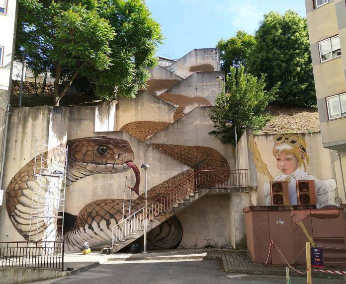 "Snake Stairs" by SFHIR in Guarda, Portugal. The mural transforms a staircase into a coiled snake, using the steps and walls to create the illusion of a slithering serpent. Beside the snake, a child dressed as an adventurer holds a staff, adding a sense of storytelling and exploration to the urban space.